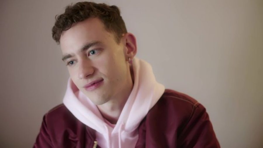 A young white man wearing a dark colours blazer over a pink hoodie. He has short, dark hair, and he looks contemplative.
